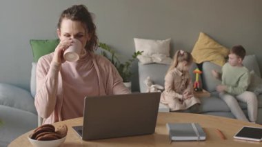 Orta yaşlı, çekici, beyaz bir kadının laptopta oturup çay içerken arka planda oynadığı bir fotoğraf.