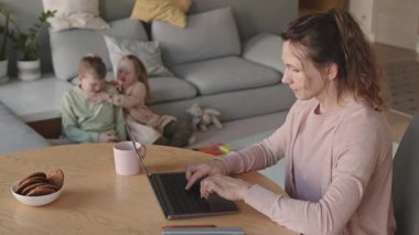 Orta yaşlı, beyaz bir kadının iki çocuğu arka planda oynarken masada oturup dizüstü bilgisayarda çalışmasının orta ölçekli fotoğrafı.