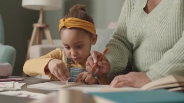 Orta boy Afro-Amerikalı bir kadın evinde oturuyor ve küçük kızına ödevlerini yaparken yardım ediyor.