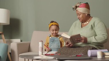 Orta yaşlı Afro-Amerikan bir kadının oturma odasında oturmuş küçük torununa kitap okurken ve defterine çizimler yaparken.