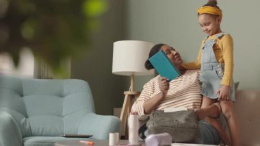 Orta boy, çekici, Afro-Amerikan bir kadının oturma odasında kanepede oturup küçük sevimli kızının okul çantasını toplamasına ve derslerine hazırlanmasına yardım ettiği bir fotoğraf.