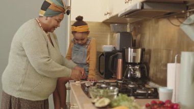 Orta boy Afro-Amerikan kadın resmi. Günlük giysiler içinde. Küçük torunu için öğle yemeği hazırlıyor.