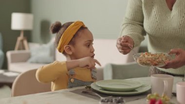 Orta boy Afro-Amerikalı bir kız yemek masasında otururken tanınmayan Afrikalı bir kadın tabağına yemek koyarken.