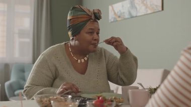 Orta yaşlı Afro-Amerikalı güzel bir kadın yemek masasında oturuyor ve tanınmayan akrabalarıyla yemek yiyor.