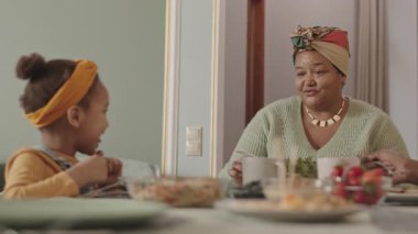 Orta boy Afro-Amerikalı kız ve büyükannesinin gündelik giysiler giyip yemek yediği bir fotoğraf.