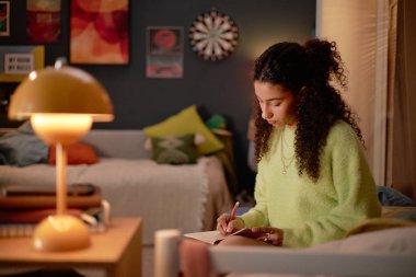 Kıvırcık saçlı, rahat, aydınlık bir odada ders çalışan, etrafı yastıklarla ve duvar süslemeleriyle çevrili genç bir kadın, özenle not defterine not alıyor.