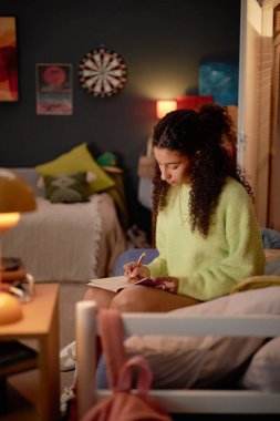 Kıvırcık saçlı, rahat bir odada oturan, yumuşak yastıklarla çevrili, rahat bir atmosfer yaratan, deftere yazı yazan genç bir kadın.