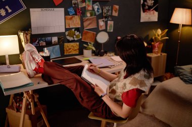 Masada çalışan Asyalı kadın rahat bir yatak odasında çeşitli dekorasyonlarla süslenmiş. Çalışma alanını aydınlatan yumuşak lamba ışığı sıcak ve davetkar bir ortam yaratıyor.