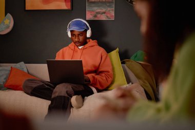 Turuncu kapüşonlu genç adam kucağında dizüstü bilgisayarla koltukta oturuyor ve kulaklık takıyor. Arkaplanda renkli yastıklarla ekrana konsantre olurken düşünceli ifade