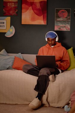 Afro-Amerikalı bir adam rahat bir odada kulaklık takmış, bilgisayar ekranında çalışırken ya da öğrenirken oturuyor.