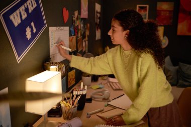 Samimi çalışma odasında takvimde yazan bir kadın. Masa lamba, defterler ve kalemler de dahil olmak üzere çeşitli öğelerle doludur.