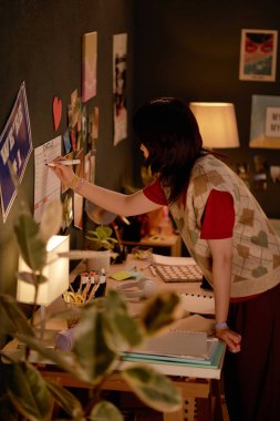 Duvarı posterler ve notlarla dekore eden kadın çalışma alanındaki kolajlara masa etrafında çeşitli malzemelerle en yeni parçaları ekleyerek