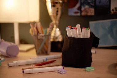 Masanın üzerinde siyah bir kapta işaretleyiciler ve arka plandaki diğer nesneler yaratıcılığa olanak sağlayan sanatsal bir çalışma alanı oluşturur.