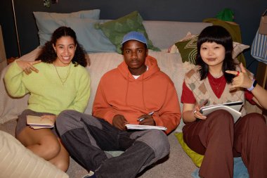 Arkadaşlar ellerinde defterler ve kalemlerle oturmuş, evde ders çalışırken kameraya gülümsüyor ve poz veriyorlar.