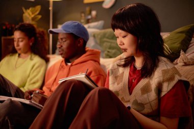 Bir grup arkadaş kanepede oturur, defterlere yazarlar ve rahat oturma odası hazırlıklarıyla meşgul olurlar. Ilık ışıklandırma rahat atmosfere katkı sağlar.