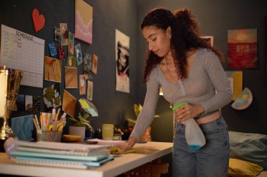 Masasını sprey sprey şişeyle temizleyen genç bir kadın fotoğraf ve duvar resimleriyle dekore edilmiş rahat bir odada, kalp şeklinde bir not ve çizelge de dahil olmak üzere.