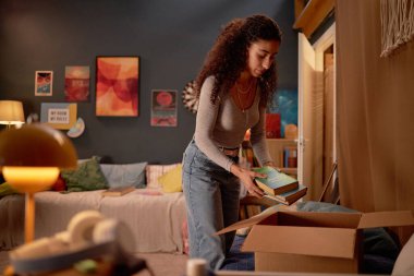 Kıvırcık saçlı genç bir kadın, kitaplarını sıcak aydınlatma ve renkli duvar dekorlarıyla dolu bir kutunun içine dikkatlice yerleştiriyor.