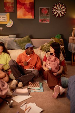 Bir grup arkadaş sıcak yurt odasında oturmuş masa oyunları ve kitapların etrafında gevşeyip sohbet ediyorlar. Canlı dekor elementleri rahat, davetkar bir atmosfer yaratır.
