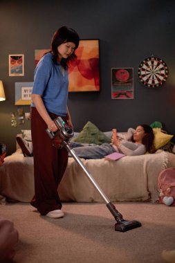 Halıda elektrikli süpürge kullanan Asyalı bir kadın. Yatak odasında, duvar süslemeleri ve yumuşak ışıklandırmalarla. Arka planda elektronik cihazla yatakta yatan kişi de yer alıyor