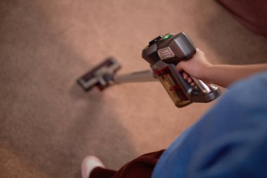 Halıyı süpürürken kablosuz süpürge tutmak, temizlik işlemine odaklanmak. Elektrikli süpürge, günlük giysiler giyen biri tarafından işletiliyor, etkin temizlik sağlıyor.