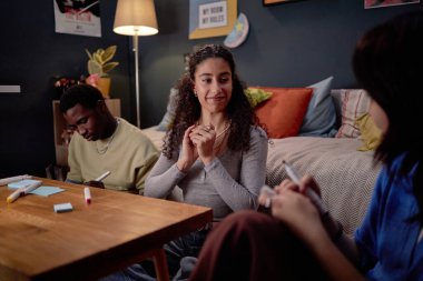 Kanepede oturmuş renkli yastıklar ve posterlerle süslenmiş, rahat oturma odasındaki ahşap sehpanın etrafında oturan iki arkadaşıyla sohbet eden gülümseyen bir kadın.