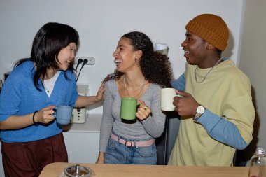 Bir grup arkadaş mutfakta durup kahve fincanlarını tutarken kahkahalarınızı paylaşarak sıcak ve dostane bir ortam yaratıyorlar.