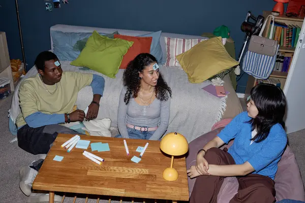 Üç arkadaş sıcak oturma odasında oyun oynamaktan keyif alıyorlar. Alınlarında yapışkan notlar var. Bu da canlı olduklarını ve birbirleriyle etkileşime geçtiklerini gösteriyor.