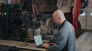 Orta yaşlı beyaz erkek tamircinin, profesyonel oto tamir atölyesinde yarı kamyonun yanında dizüstü bilgisayarda çalışırken orta ölçekli görüntüsü.