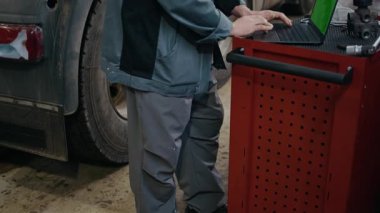 Orta yaşlı beyaz erkek tamircinin yarı kamyon tamirhanesinin önünde dizüstü bilgisayarla çalışırken görüntüsünü kaldır.