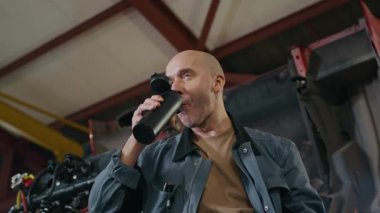 Düşük açılı, orta yaşlı, beyaz işçinin garajda termos şişesinden kahve içerken ki görüntüsü.