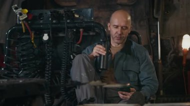 Orta yaşlı Kafkasyalı tamircinin cep telefonunu kaydırırken termos şişesinden kahve içerken, oto tamirhanesinde kahve molası verirken görüntüsü.