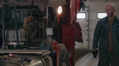 Orta yaşlı beyaz erkek tamircinin el kamerası görüntüsü oto tamirhanesinde yarı kamyona yaklaşıp termos şişesinden kahve yudumlarken görüntüyü kontrol ediyor.