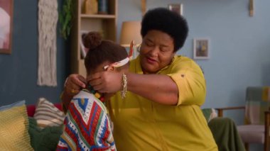 Afrikalı Amerikalı kız büyükannesiyle kolye ve bilezik deniyor, rahat oturma odasında kanepede oturuyor.