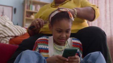 Tanımlanamayan Afrikalı Amerikalı büyükannenin, akıllı telefondan oyun oynarken torununun saçını yaptığı düşük açılı çekim.