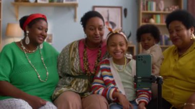 Üç kadın, küçük çocuk ve bebek, renkli kıyafetler ve aksesuarlar giyip, kanepede oturuyorlar, rahat oturma odasındaki tripodda akıllı telefona gülümsüyorlar.