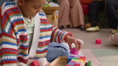 Afrika kökenli Amerikalı bir kız yerde renkli bloklarla oynarken, bir kadın kucağında bebek, konuşuyor ve gülümsüyor, rahat bir oturma odasında kanepede oturuyor.