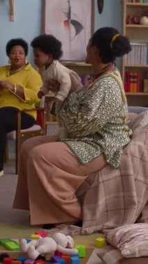 Dikey çekimde, Afrikalı Amerikalı büyükannenin çocuklarıyla oturma odasında örgü örüp konuştuğu fotoğraf.