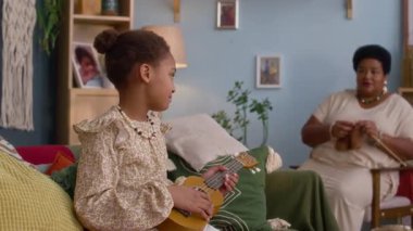 Büyükannesi onunla konuşurken ve oturma odasında örgü örerken kanepede oturup ukulele çalan Afrikalı Amerikalı bir kız.