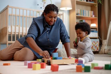Siyah ırkın genç annesi küçük bir bebekle oynuyor halının üzerinde oturmuş yatak odasındaki ahşap bloklarla renkli yapılar inşa ediyor.