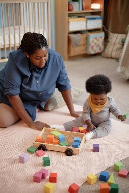 Dikey çekimde, siyah anne küçük bir bebekle vakit geçiriyor. Renkli yapı taşları ve ahşap arabalarla oynuyor. Yatak odasındaki halıda oturuyor.