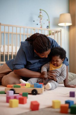 Siyah annenin, mutsuz küçük bebeği rahatlatırken yatak odasındaki renkli yapı taşlarıyla oynarken iyi ağladığı dikey bir fotoğraf.