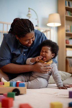 Dikey çekimde, siyah anne ağlayan küçük kızını rahatlatırken yatak odasındaki yapı taşlarıyla dolu bir halının üzerinde otururken.