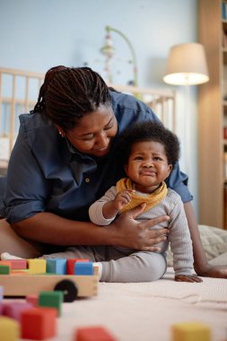 Dikey çekimde, siyah bir anne, küçük bir çocuğu rahatlatıyor. Kızına nazikçe sarılıyor. Yatak odasında çeşitli oyuncaklarla dolu bir halının üzerinde oturuyor.