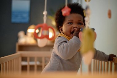 Çocukların yatak odasındaki renkli müzik cep telefonlarıyla ahşap bir beşikte dikilirken orta boy siyah bebek diş çıkarma oyuncağını ısırıyor.