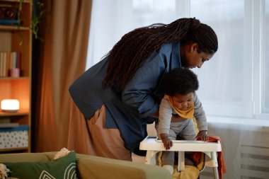 Siyahi bir annenin, küçük kızını sandalyeye oturtup bebeğini akşam yemeğine hazırladığı orta boy bir fotoğraf.