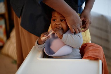 Çocuğunu akşam yemeğine hazırlayan bebeğe silikon önlük takan şefkatli siyah annenin elleri. Bu arada çocuk da sıcak ışıklarla mutfakta yüksek sandalyede oturuyor.