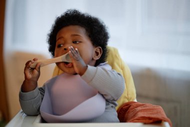Yemek sırasında silikon önlük takan siyah bir kızın sıcak bir tonla mutfakta yüksek sandalyede oturduğu orta boy bir fotoğraf.