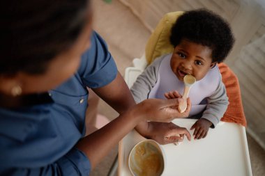Güzel siyah kız bebek yüksek sandalyede oturur ve kameraya bakar, annesi mutfakta kahvaltıda kaşıkla çocuğu beslerken.