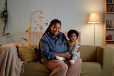 Küçük kızını kucağında taşıyan mutlu siyah anne pozu veren orta boy bir fotoğraf. O sırada küçük kız çocuğuyla rahat bir yatak odasında kanepede oturuyor.
