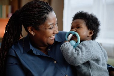 Orta ölçekli, neşeli, siyah bir kadın, elinde mutlu bir bebeği tutarken çocuk diş büyütme oyuncağını ısırıyor.